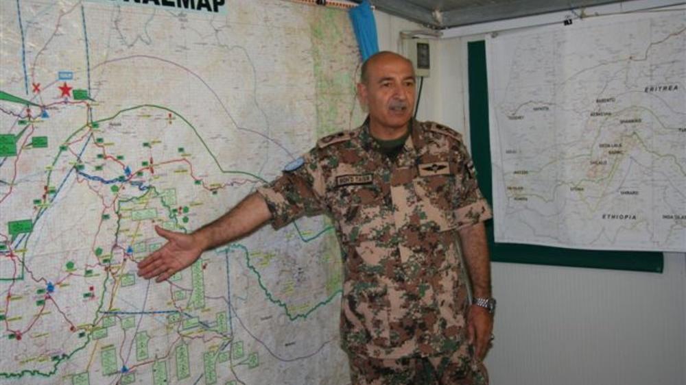 A man in military uniform standing in front of a large map indicating a section of the map with an open hand.