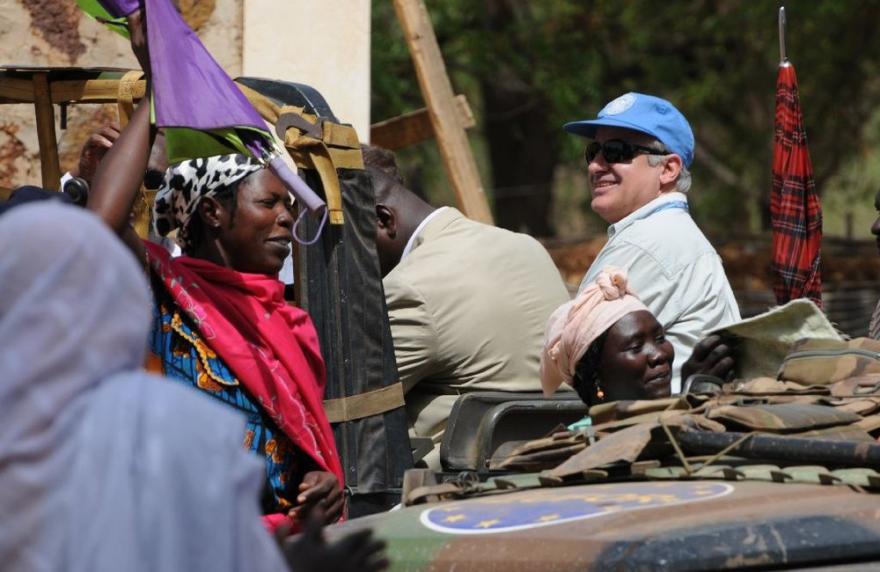 People gather near a military vehicle with UN markings; colorful fabrics and equipment are visible in the scene.